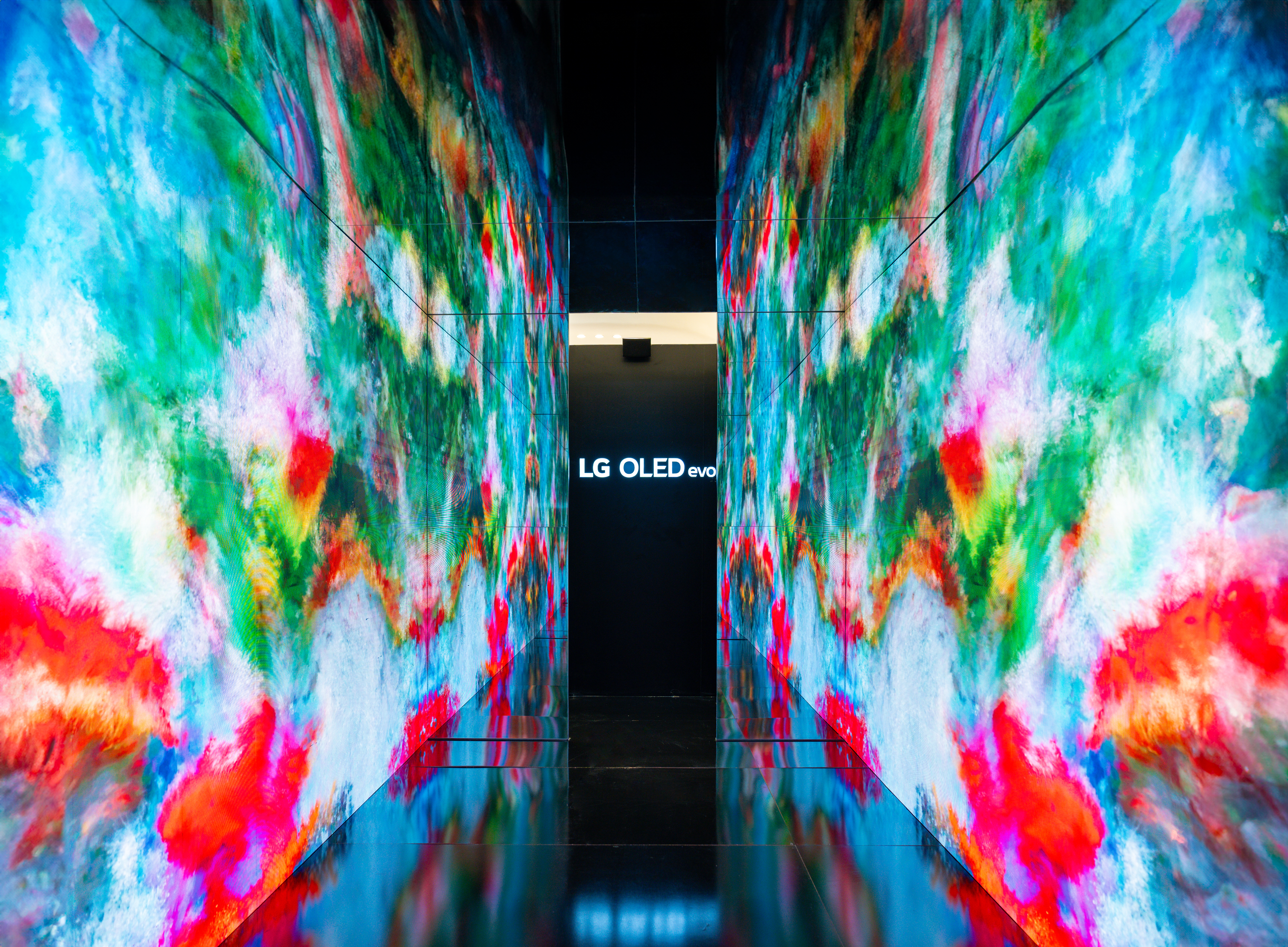 The immersive walls displaying an alternate generative installation. The screens show textured patterns in cool tones of blue, green, and red, reflecting across the dark floor toward the central signage.